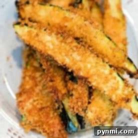 Golden, crispy air fryer zucchini fries stacked on a white plate, coated in breadcrumbs.
