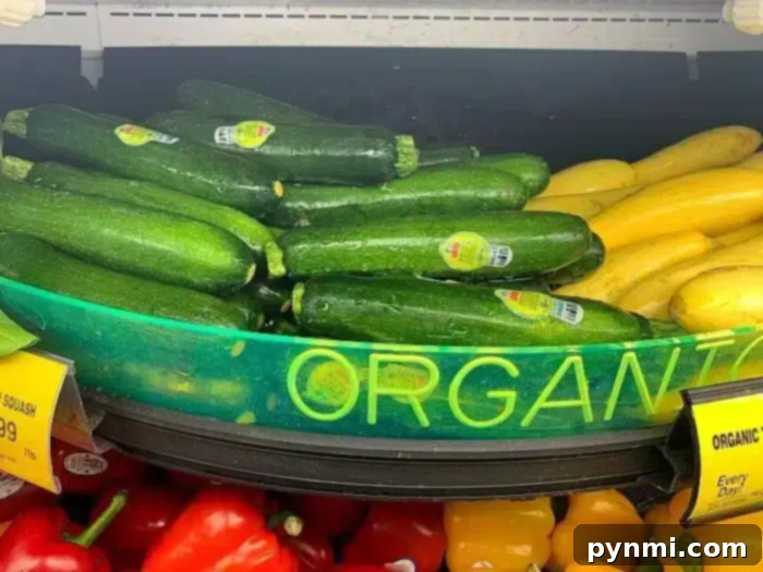 A grocery store display features green zucchini with produce stickers and yellow squash, perfect for making air fryer zucchini fries. A partially visible yellow sign reads "ORGANIC." Red bell peppers are visible in the bottom of the frame.