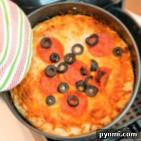 A baked air fryer pizza with a golden cheese crust, topped with sliced pepperoni and black olives, is being held in a pan using a striped oven mitt.