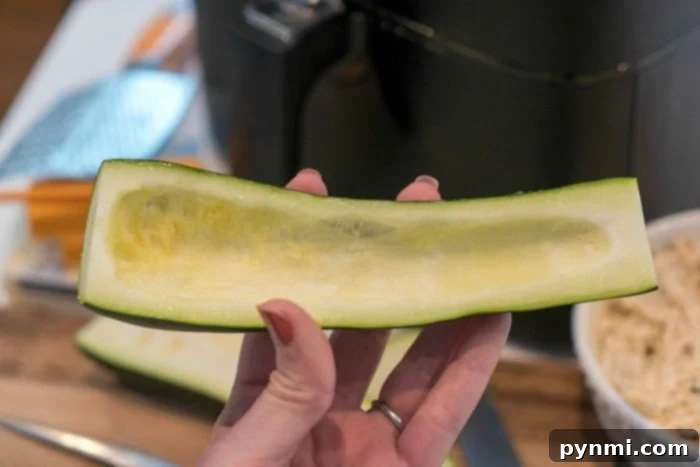 Two hands scooping out the seeds and flesh from a halved zucchini using a spoon, preparing it for filling.