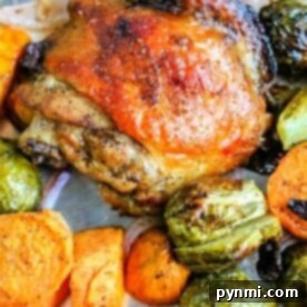 Sheet pan chicken thighs with charred skin, nestled among baked sweet potatoes, Brussels sprouts, and red onion on a baking sheet.