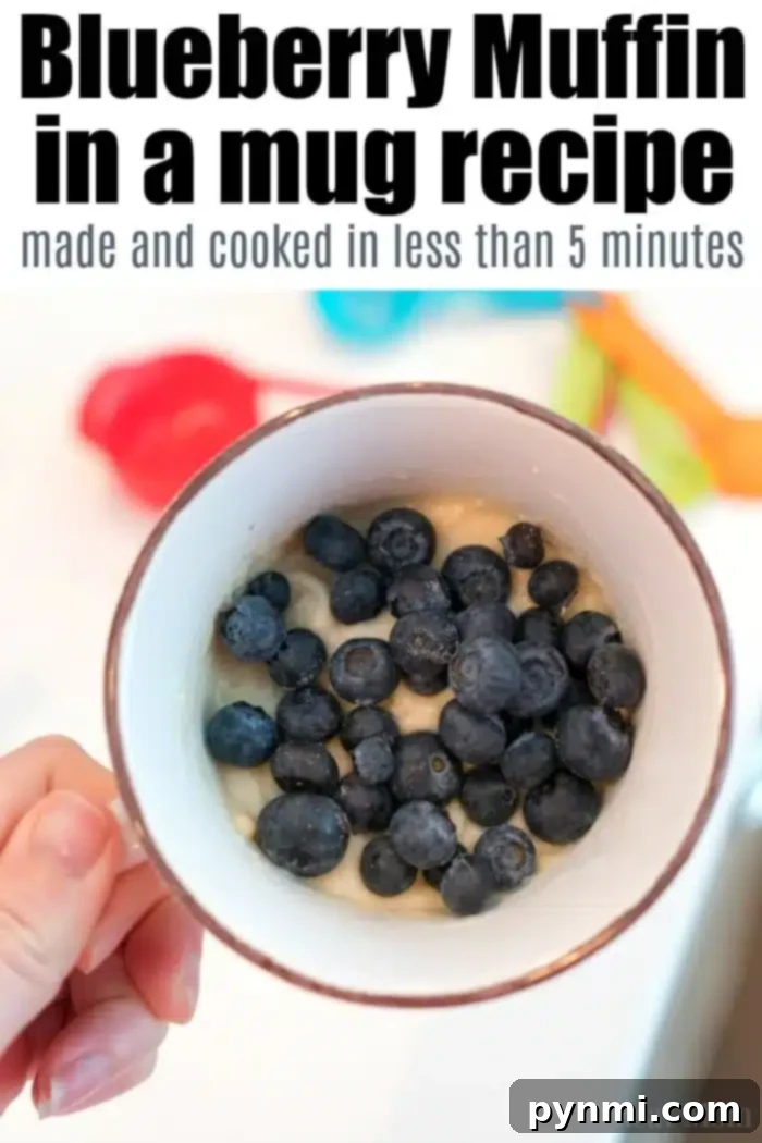 A hand holds a mug packed with blueberry muffin in a mug batter, topped generously with fresh blueberries. The white mug with a brown rim promises delight. The text above proclaims, "Blueberry Muffin in a Mug made and cooked in under 5 minutes," while kitchen items softly fade into the background.