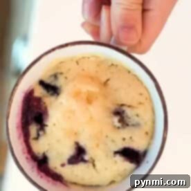 A hand holding a mug filled with a baked blueberry muffin in a mug cake, revealing juicy blueberries inside.
