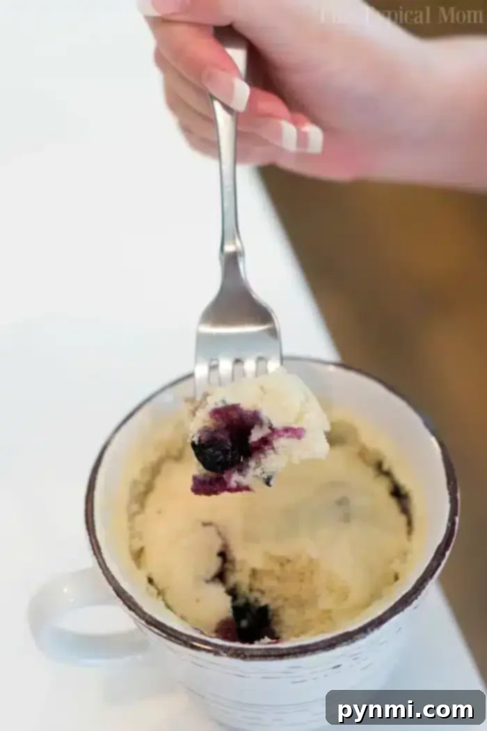 A hand holding a fork with a piece of blueberry muffin in a mug above a white ceramic mug filled with the cake. The mug is placed on a white surface.