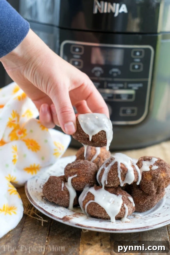 Easy air fryer donut recipe using cinnamon rolls and cinnamon and sugar! Soft on the inside and crispy outsides, make these in your Ninja Foodi tonight! #airfryer #airfryerrecipe #donuts #cinnamonrolls #ninjafoodi #ninjafoodirecipes #dessert A hand holds an iced air fryer donut ball made with cinnamon rolls, with a Ninja Foodi air fryer in the background.