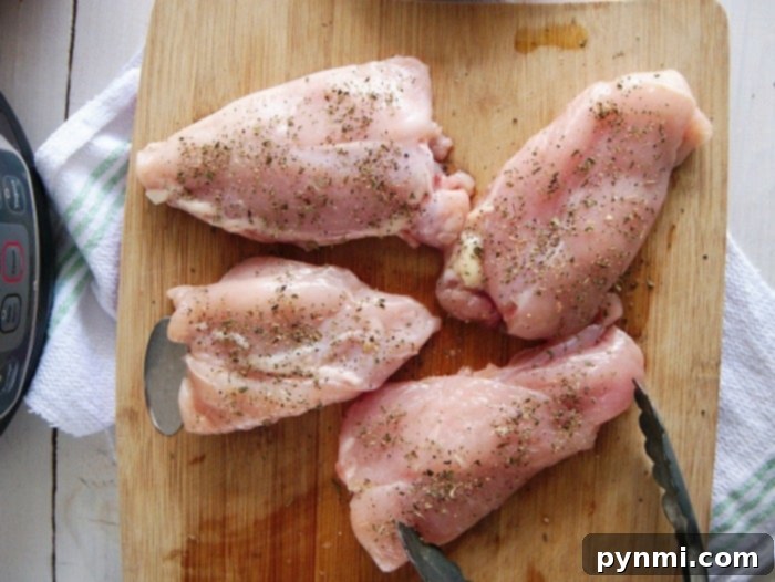 Four seasoned raw chicken thighs, perfect for a crockpot lemon pepper chicken recipe, rest on a wooden cutting board alongside a trusty pair of tongs, ready for the slow cooker.
