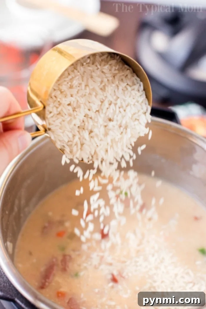 Sausage and rice in an Instant Pot is so good! A cheesy casserole packed with smoked sausage, tender rice and vegetables cooked all in one pot in no time at all. #instantpot #instantpotrecipes #sausage #rice #casserole #cheesy #pressurecooker #ninjafoodi Pouring uncooked rice from a measuring cup into an Instant Pot with simmering sausage and rice casserole ingredients.