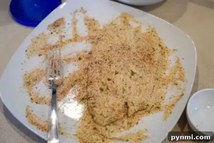 Golden Crispy Air Fryer Tilapia 5 A piece of air fryer tilapia is covered in a seasoned breadcrumb mixture on a white plate, with a silver fork resting on the side.