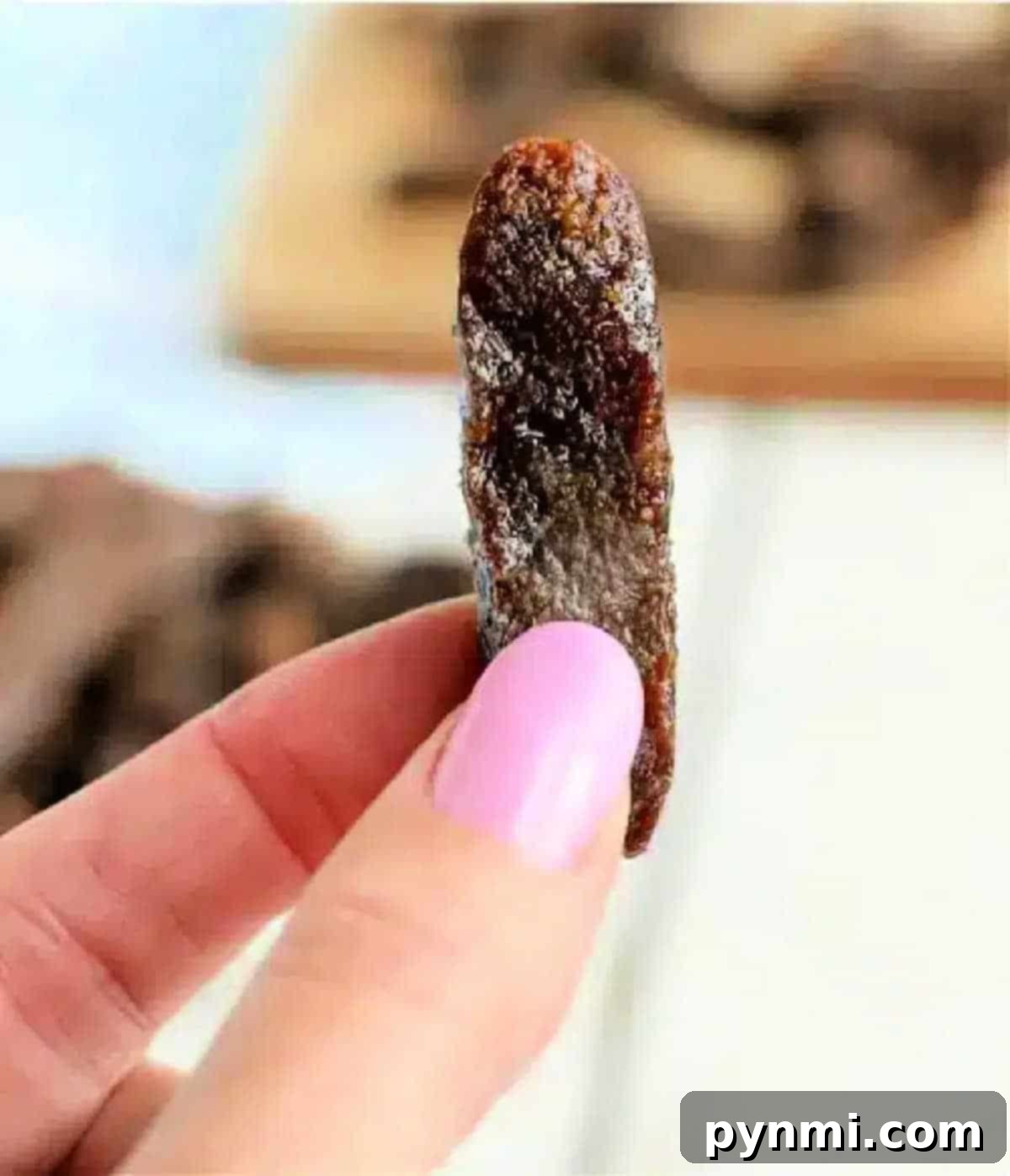 A hand with pink nail polish holds a single dried tamarind piece close to the camera, reminiscent of air fryer beef jerky in its texture and appearance.