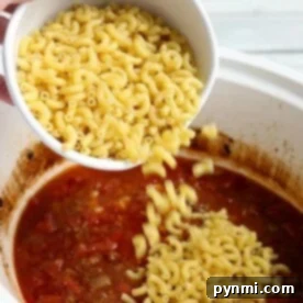 Pouring uncooked elbow macaroni into a simmering pot of crockpot chili mac with a rich tomato base adds hearty texture and delicious flavor.