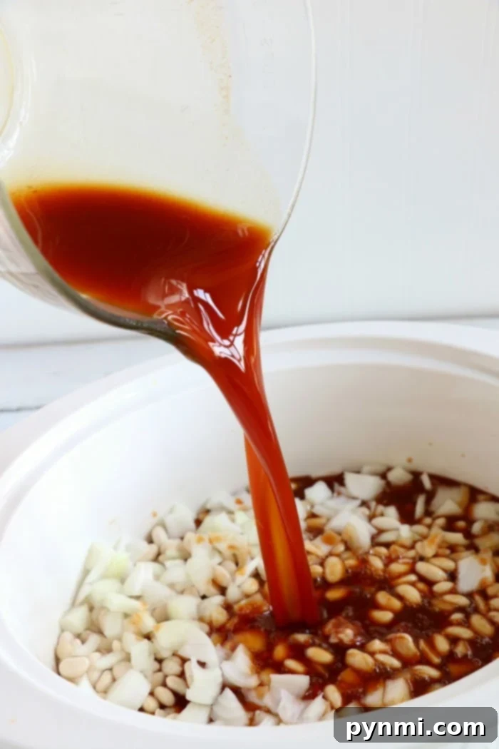 Homemade Crockpot baked beans uses dry beans, and so much better than canned! You'll never buy them at the store again after trying these. #crockpotrecipes #crockpotbakedbeans #bakedbeans #slowcooker #homemadebakedbeans #homemade #sidedsih Pouring sauce into a slow cooker with chopped onions and white beans creates the perfect base for a homemade crockpot baked beans recipe.