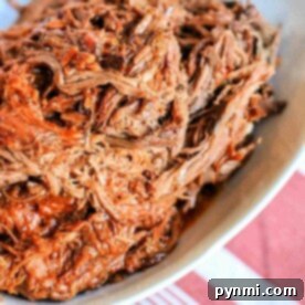 Crockpot BBQ beef, tender and flavorful, sits shredded in a white dish on a table with a red and white striped cloth.