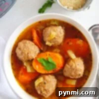 A white bowl contains instant pot meatball soup with sliced carrots and celery, garnished with a parsley leaf. A spoon is on the side, and a small bowl with grated cheese is in the background on a white surface.