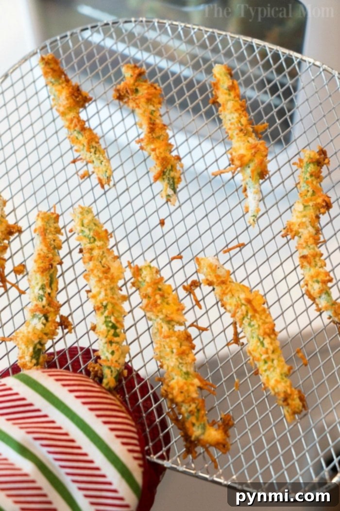 Air fryer green beans with parmesan are a great snack or side dish with any meal. Crispy and crunchy vegetables covered in Panko bread crumbs and seasoning. #airfryerrecipes #airfryer #greenbeans #greenbeanfries #airfryergreenbeanfries #airfryergreenbeans #thetypicalmom A wire rack with crispy, golden-brown green bean fries held by a person with an oven mitt.