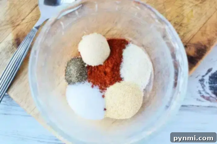 A clear glass bowl contains piles of spices perfect for seasoning the best baked pork chops: black pepper, paprika, salt, garlic powder, onion powder, and brown sugar. Nearby, a fork and knife rest on a wooden cutting board.