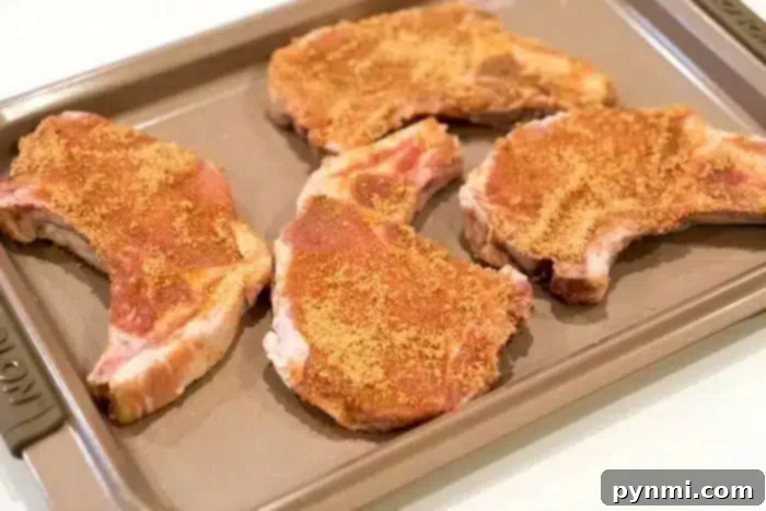Four seasoned pork chops arranged on a baking sheet, ready for cooking. The chops, promising to be the best baked pork chops you've tasted, are sprinkled with a brown spice mix. The baking sheet is placed on a light-colored surface.