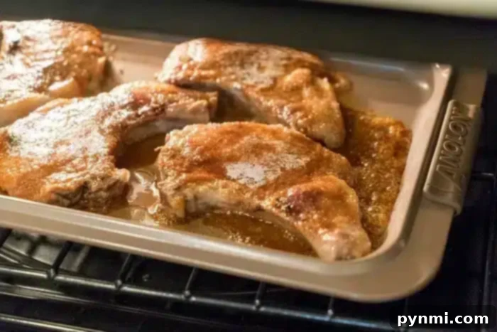 Baked pork chops on a metal baking tray inside an oven, showcasing the best baked pork chops. Coated with a golden brown seasoning and surrounded by their juices, the tray features a handle with the brand name "Anolon" visible.