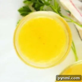 Top view of a glass jar filled with yellow liquid, resembling chicken broth, on a white background with a parsley sprig nearby.