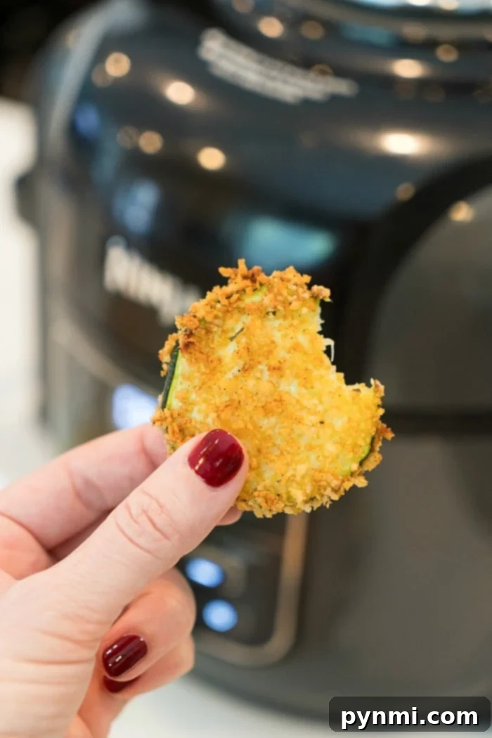 A hand holding a bite of crispy breaded zucchini in front of an air fryer, with red-painted nails, showcasing the perfect snack alongside freshly made air fryer zucchini chips, highlighting their irresistible texture.