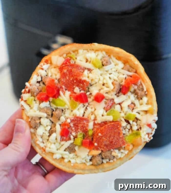 Air Fry Your Frozen Pizza to Perfection 2 A hand holds a small, round frozen pizza, topped with diced green and red bell peppers, pepperoni slices, sausage crumbles, and shredded cheese. In the background is a black appliance—perfect for cooking frozen pizza in an air fryer.