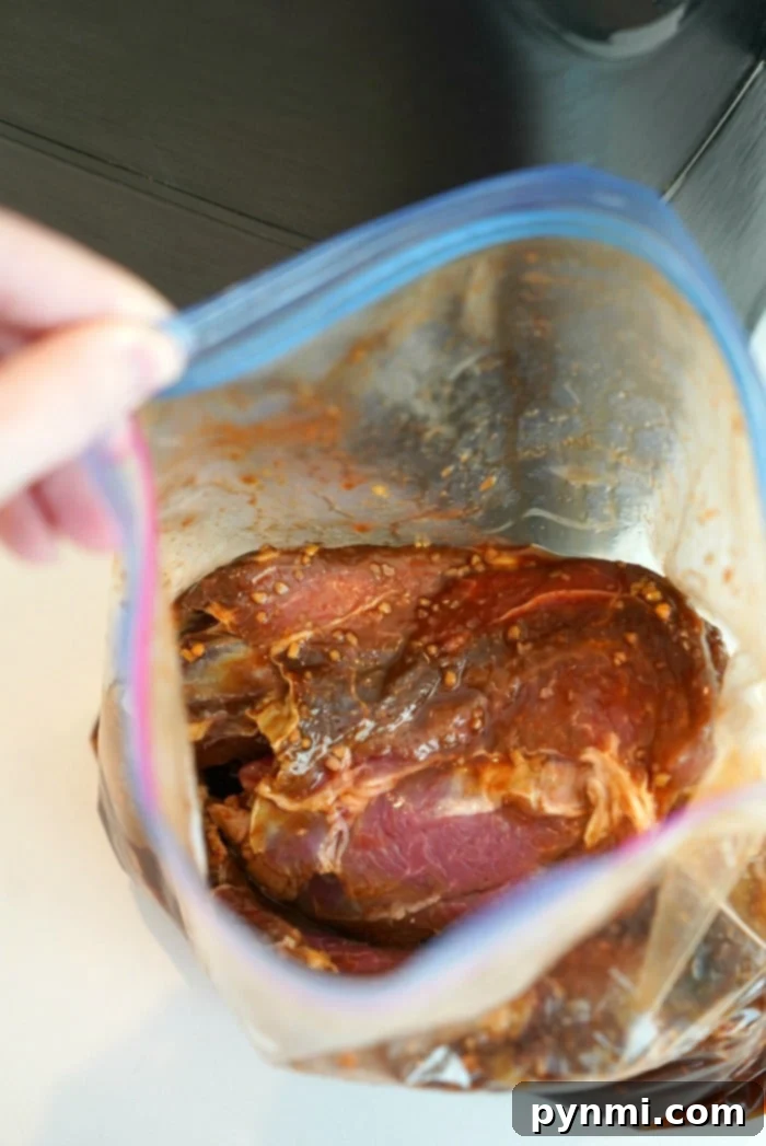 A hand holds an open plastic bag containing raw beef roast pieces generously coated in a vibrant red teriyaki marinade, ready for air frying. The rich color of the marinade and visible garlic pieces suggest deep flavor.