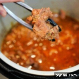 A close-up shows a hand holding metal tongs with a piece of succulent carnitas above a pot brimming with meat in a rich, red sauce. The pot appears to be part of a pressure cooker, ensuring the flavors meld perfectly.