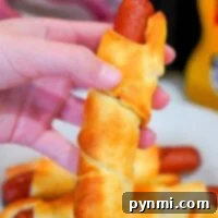 A hand holds a baked crescent roll wrapped around a hot dog, reminiscent of classic air fryer pigs in a blanket. In the background, more wrapped hot dogs rest on a white plate. A bottle of mustard is partially visible to the right.