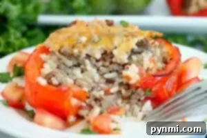 Perfect Air Fryer Stuffed Peppers 10 A stuffed red bell pepper, blanched to perfection, sits on a white plate filled with a savory mix of ground meat and rice, topped with melted cheese. Diced tomatoes and chopped herbs are artfully scattered around the pepper, while a fork rests beside it.