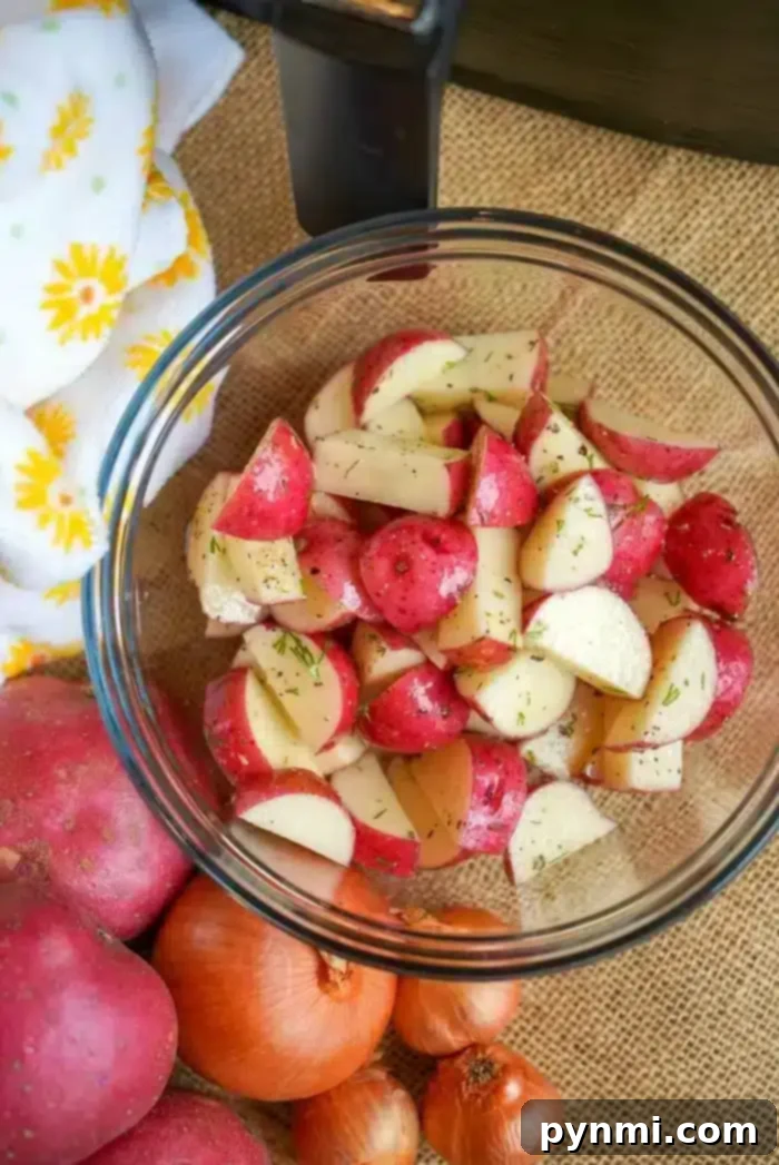 A glass bowl filled with chopped red potatoes seasoned with herbs, ready for the air fryer. Surrounded by whole red potatoes, onions, and a decorative cloth.