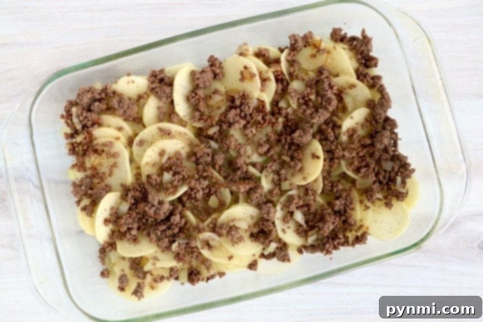 Layers of ground beef and potatoes in a baking dish before adding cheese and sauce.