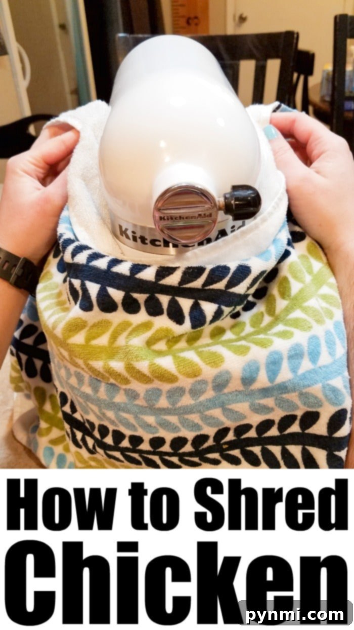 A stand mixer with a paddle attachment vigorously shredding cooked chicken in a bowl, covered by a hand towel to prevent splattering.
