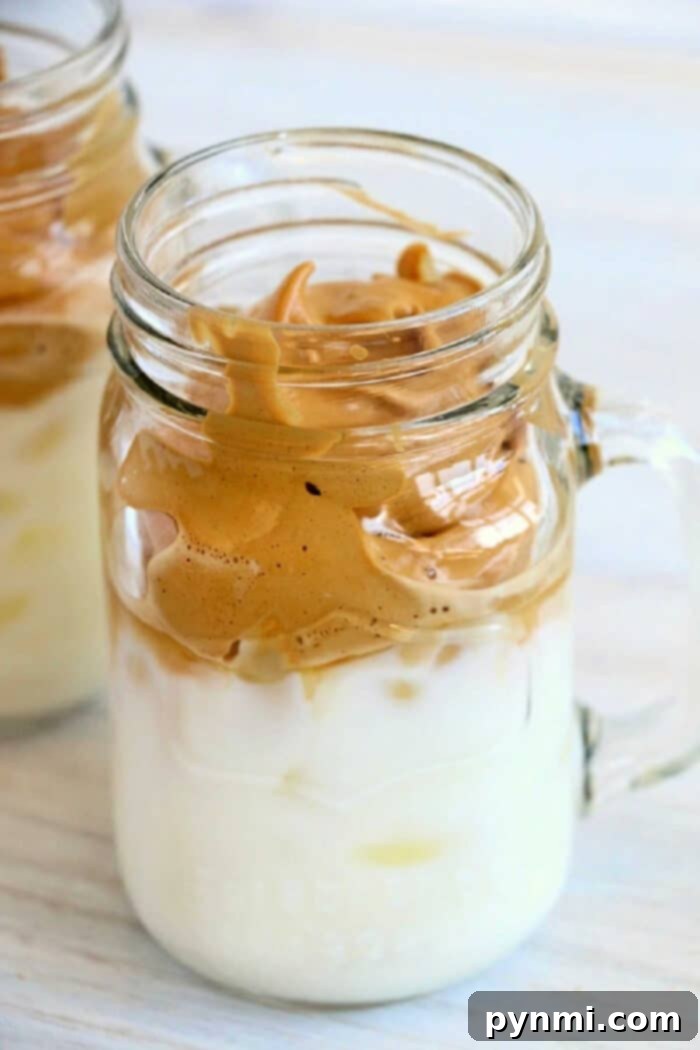 Try this 3 ingredient trendy Dalgona whipped coffee recipe! EASY to make this popular TikTok drink everyone is going crazy over. I bet you'll love it too. Dalgona Whipped Coffee: A close-up of the frothy coffee topping over milk in a clear glass, garnished with a sprinkle of coffee powder.