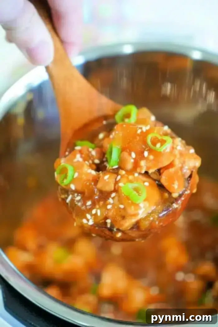 Sticky Sweet Instant Pot Korean Chicken 2 A close-up of a wooden spoon holding a serving of an Instant Pot Korean chicken dish with diced vegetables in a thick sauce. The dish is garnished with sesame seeds and sliced green onions. The background shows a pot containing the rest of the flavorful meal.