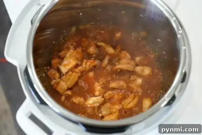 Sticky Sweet Instant Pot Korean Chicken 6 A close-up view of an Instant Pot containing chicken pieces simmering in a red tomato-based sauce. The dish, possibly Korean chicken, appears to be in the process of cooking, with visible steam and some chopped onions mixed in.
