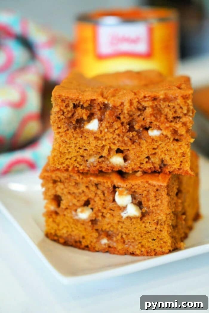 Two delicious pumpkin blondies with white chocolate chips are stacked invitingly on a pristine white plate, set against a vibrant, colorful cloth in the background.