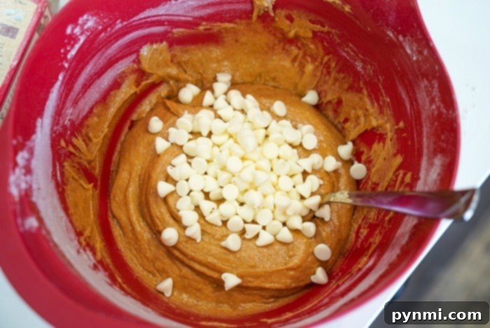 A red mixing bowl brimming with dough and white chocolate chips promises delightful pumpkin blondies, while a metal spoon rests inside, ready for action.