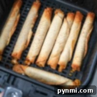 An air fryer basket contains several frozen taquitos arranged in neat rows, now transformed into crispy delights. Each taquito is golden brown with some browned edges, and a bit of cheese has melted out from the ends.