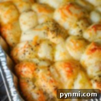 A close-up of a baked dish in a foil tray, showcasing golden-brown smoked bread covered with melted cheese and sprinkled with herbs. The bread appears fluffy and slightly crispy on top, with unevenly distributed cheese and herbs.