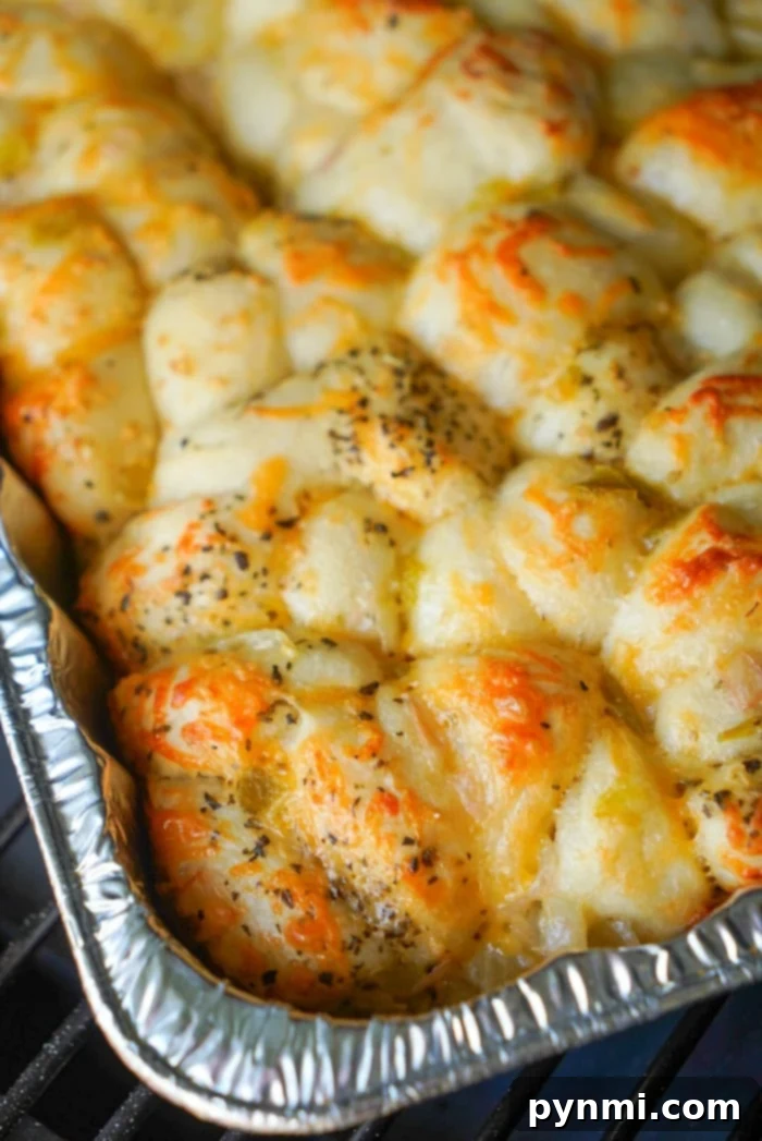 A close-up of a baked dish in a foil tray, showcasing golden-brown smoked bread covered with melted cheese and sprinkled with herbs. The bread appears fluffy and slightly crispy on top, with unevenly distributed cheese and herbs.