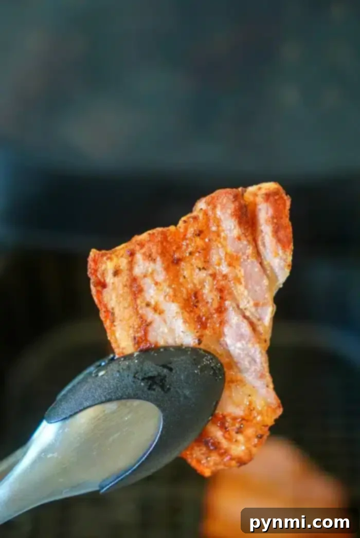 A piece of seasoned and cooked air fryer pork belly is being held by a pair of metal tongs. The background is slightly blurred and appears to be an air fryer. The pork belly is visibly crispy and browned around the edges.