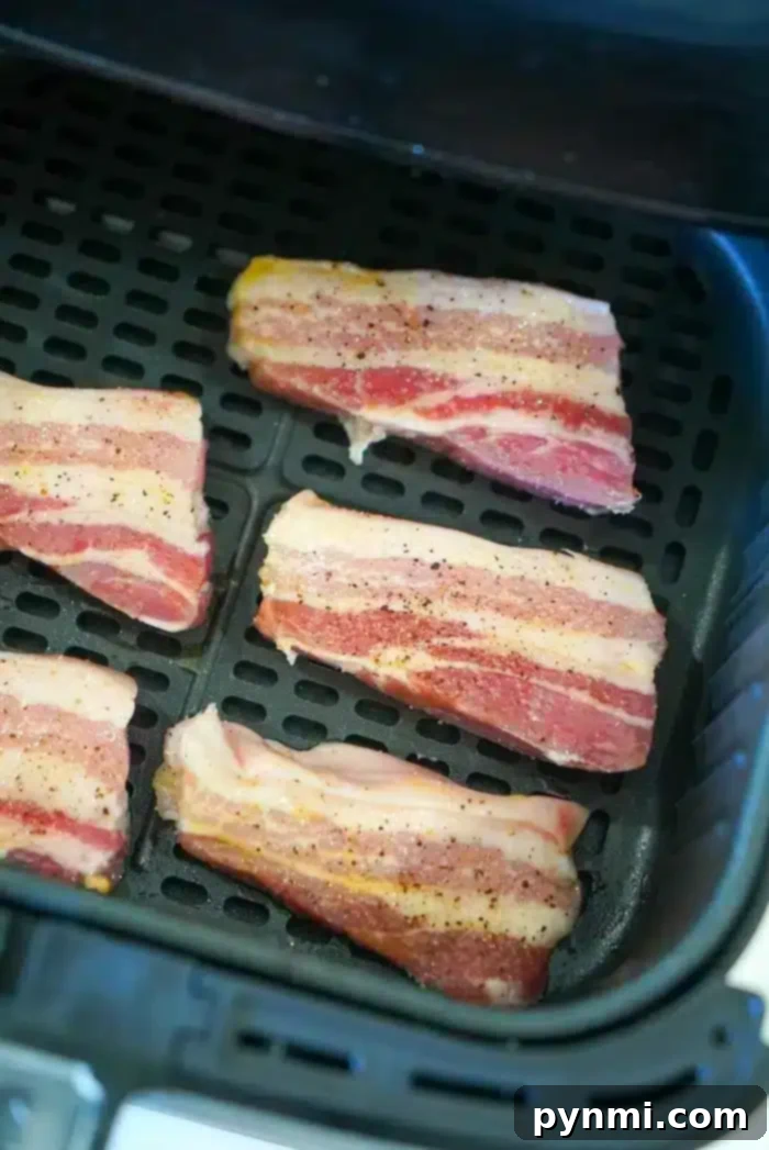 Six strips of raw bacon are arranged on the tray inside an air fryer, ready to be cooked. The bacon strips, seasoned with black pepper and salt, resemble thin pieces of pork belly. The air fryer tray has a grid pattern for even cooking.