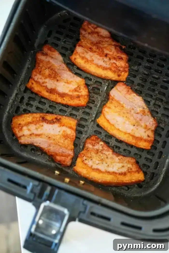 Six pieces of pork belly bacon, partially cooked with browned edges, are arranged inside an air fryer basket. The basket has a black grid-like base and sits on a white surface.