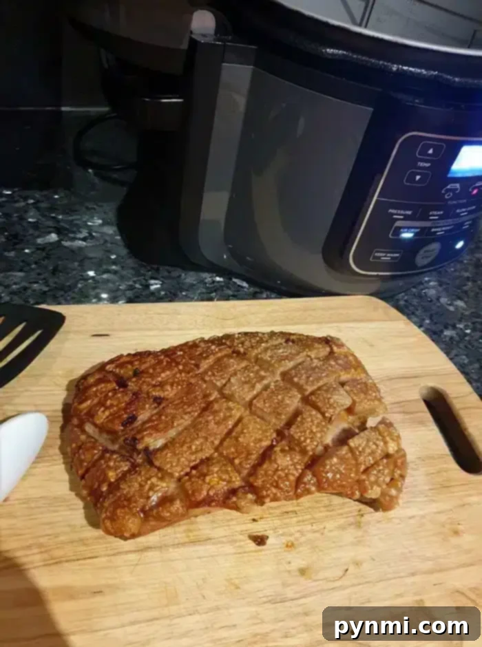 A wooden cutting board holds a piece of cooked pork belly with crispy, cross-hatched crackling. A spatula handle is visible on the left, and a slow cooker with digital controls sits in the background on a dark kitchen countertop, highlighting the results of perfectly air-fried pork belly.