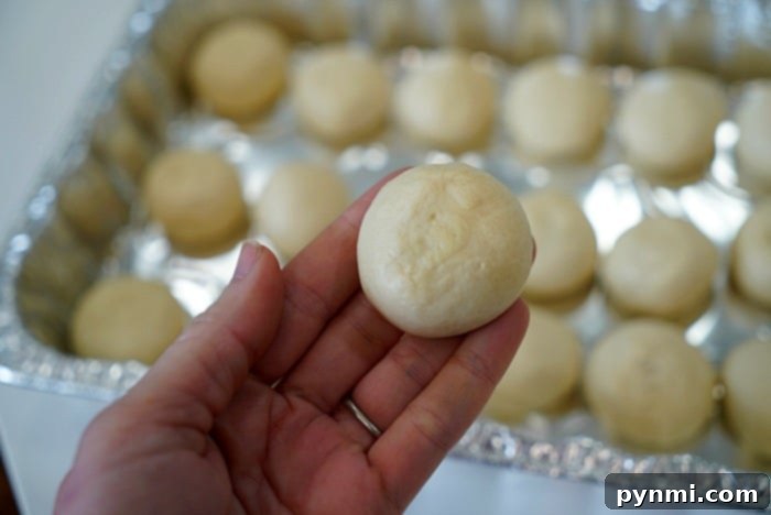 A person delicately holds a small, round dough ball above a tray of similar dough balls, each destined to become a piece of aromatic smoked bread.