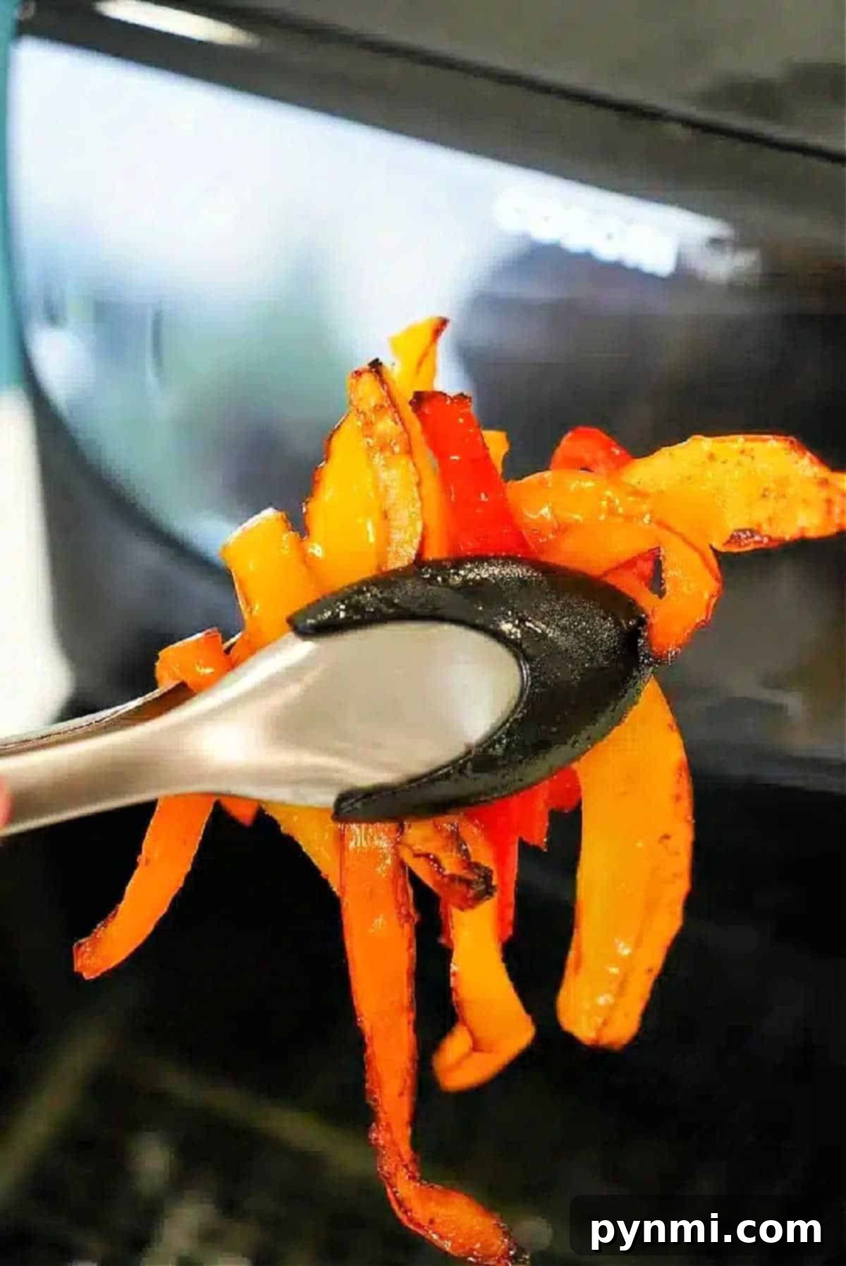 Tongs holding cooked orange and red air fryer peppers in front of an air fryer.