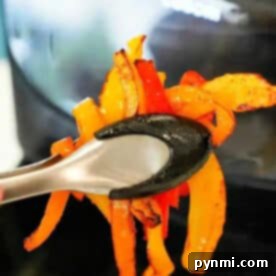 Tongs holding cooked orange and red air fryer peppers in front of an air fryer.