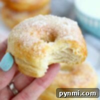 A hand holds a bitten, sugar-dusted cronut, reminiscent of a delightful fusion between biscuit and donut, with more cronuts stacked enticingly in the background.