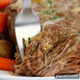 Close-up of a fork shredding tender pot roast made in a Ninja Foodi, with carrots and potatoes in the background.