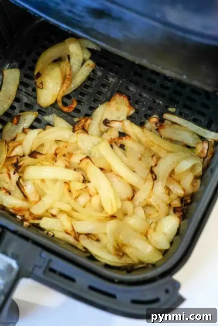 Crispy Air-Fried Onions 2 Sliced onions are shown cooking in an air fryer basket, partially browned and in the process of caramelizing. The air fryer is open to display the air fryer onions inside.