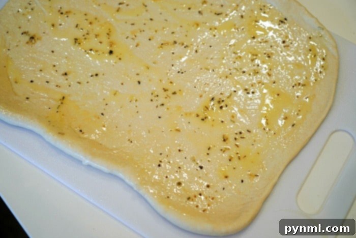 Pizza Pinwheels 4 A sheet of uncooked thawed from frozen pizza dough is evenly spread with olive oil and sprinkled with herbs, positioned on a white cutting board.